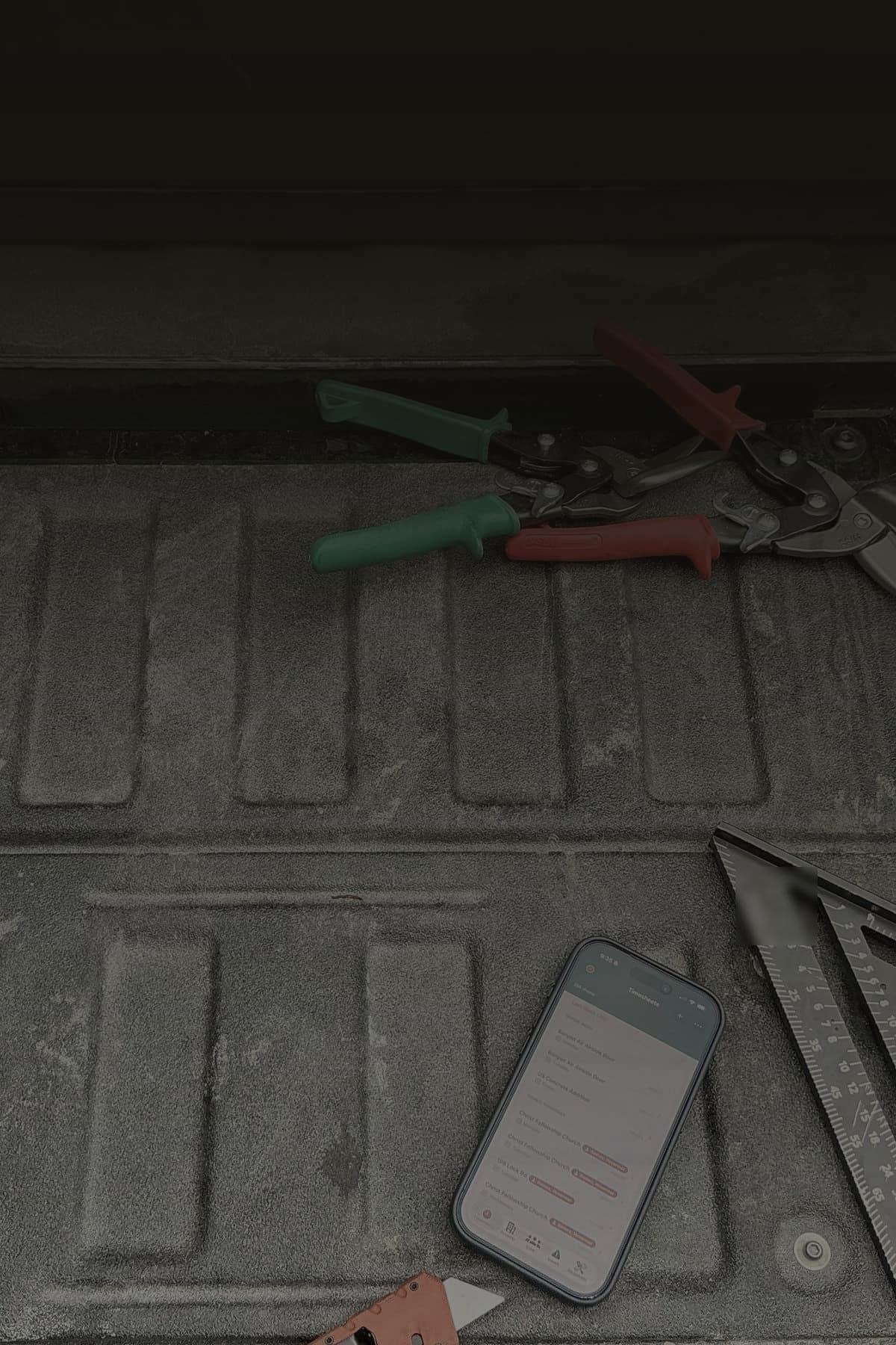 Jobsite tools and an iPhone running a SiteSuite app laid out on a truck tailgate — snips, a rafter square, a level, and a wrench.